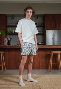 Hvit t-skjorte med kort arm, mønstrede lys grønne og beige shorts med virvlende design, hvite crew-sokker, står på vevet teppe på et kjøkken.