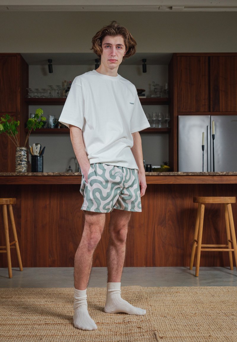 Hvit t-skjorte med kort arm, mønstrede lys grønne og beige shorts med virvlende design, hvite crew-sokker, står på vevet teppe på et kjøkken.