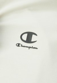 Weißer Stoff mit einem strukturierten Rippmuster, versehen mit einem schwarzen Champion-Logo und Schriftzug. Das Design ist minimalistisch und zeitgenössisch.