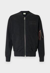 Chaqueta bomber negra hecha de algodón. Cuenta con una cremallera frontal, bolsillos laterales y un bolsillo con cremallera en la manga con una lengüeta de tirador en rojo y blanco.