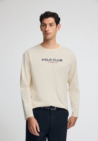 Camicia a maniche lunghe beige chiaro con la scritta "POLO CLUB" stampata in blu navy e rosso. Tessuto morbido, colletto girocollo e vestibilità comoda, abbinata a pantaloni scuri.