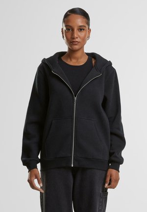 Kvinne med bakglattet hår iført en svart hettegenser med glidelås og matchende svarte bukser, stående mot en ensfarget grå bakgrunn.