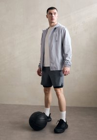 Lichtgrijs jack met ritssluiting over een wit T-shirt, gecombineerd met donkere korte broek en zwarte sneakers, staand naast een zwarte basketbal op een neutrale vloer.