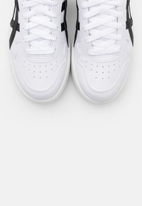 Paire de baskets blanches avec des bandes latérales noires, zone des orteils perforée et lacets blancs, vues de dessus sur fond blanc.