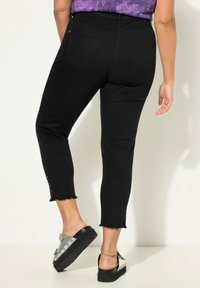 Jeans negros cortos con dobladillo deshilachado, que cuentan con bolsillos traseros. Llevados con sandalias plateadas adornadas. Textura suave y diseño ajustado.