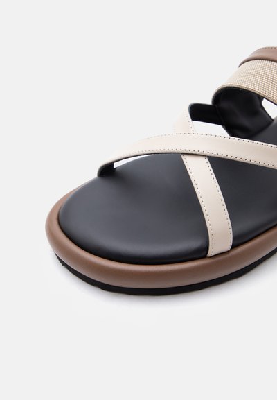 Πέδιλο με ανοιχτά δάχτυλα, μαύρο πάτο, καφέ σόλα και μπεζ σταυρωτά λουράκια από ραμμένο υλικό.