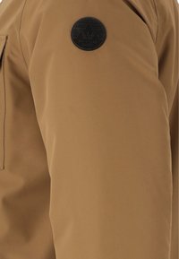 Brun ytterkläder med en slät textur, med ett svart cirkulärt logomärke märkta "WHISTLER" på vänster ärm.