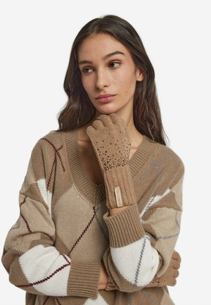 Gants sans doigts en tricot beige avec des accents texturés et des motifs découpés. Portés avec un pull beige présentant un motif en losange de différentes couleurs.