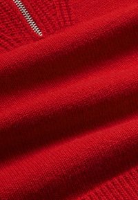 Détail d'un pull en tricot rouge mettant en avant une texture lisse, un ourlet côtelé et une fermeture éclair en argent le long de l'encolure.