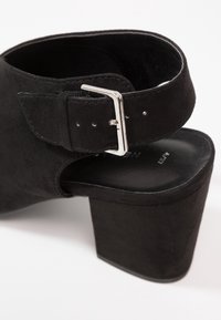 Sandale à talon haut en daim noir avec une large base, une sangle à boucle réglable et un intérieur lisse. Présente un design à découpe arrondie.