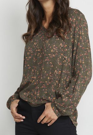 Mujer que lleva una blusa de manga larga verde oliva con un pequeño estampado floral y pantalones negros, con las manos apoyadas cerca de los bolsillos.