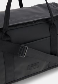 Borsa a duffle nera, strutturata e testurizzata, con forma rettangolare, manici robusti e chiusura a zip. Presenta una tracolla con fibbia e un patch in pelle con il logo.