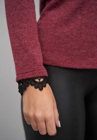Bordeauxroter Pullover mit figurbetontem Schnitt; verfügt über eine schwarze Spitzenmanschette mit floralem Muster. Hand ruht auf einer schwarzen Leggings.