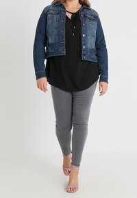 Denim-takki, jossa on sininen kangas ja napitus, yhdistetty mustaan yläosaan, harmaisiin kapeisiin farkkuihin ja vaaleanliiloihin sandaaleihin.