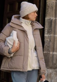 Giacca imbottita beige con cappuccio, indossata sopra un top in pile bianco. Il modello tiene in mano un oggetto bianco e soffice e indossa un berretto di lana beige.