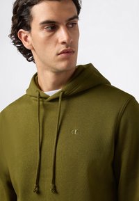 Mladý muž s kučeravými vlasmi, oblečený v olivovo zelenej mikine s kapucňou a malým vyšitým logom, hľadí doľava pred jednoduchým pozadím.