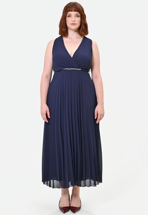 Donna con capelli rossi che indossa un vestito plissettato blu navy senza maniche con dettaglio di perle in vita, in piedi davanti a uno sfondo chiaro e semplice.