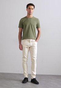 Olivově zelené bavlněné tričko, slim-fit smetanové džíny a černé tenisky. Model stojí s rukama v kapsách na neutrálním pozadí.