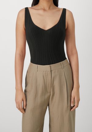 Vrouw draagt een mouwloos, zwart geribd topje met V-hals, ingestopt in hooggetailleerde beige geplooide broek.