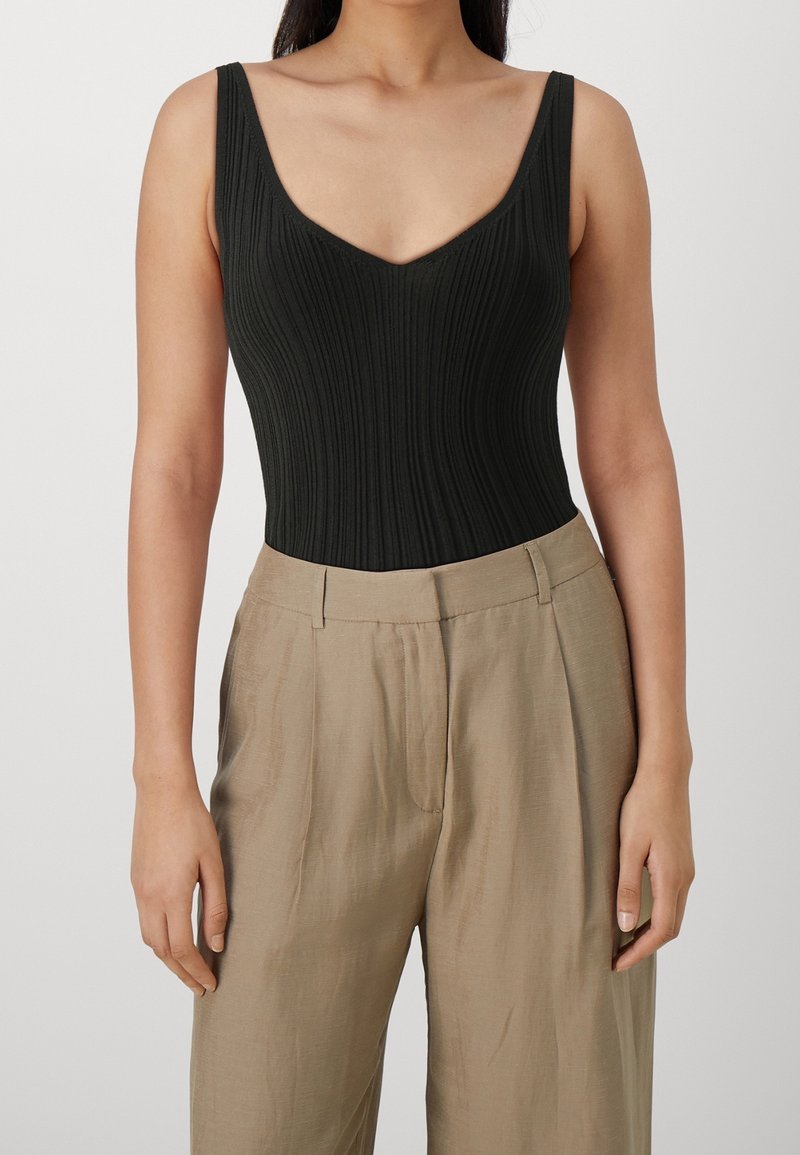 Femme portant un haut noir côtelé sans manches à col en V, rentré dans un pantalon beige plissé taille haute.