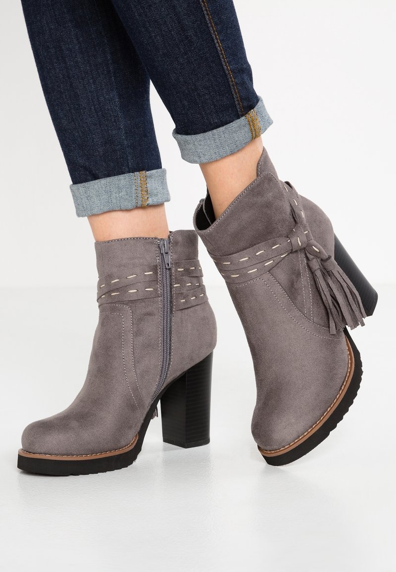 Bottes en daim gris montant à la cheville avec talons blocs, fermetures éclair latérales, bandes décoratives cousues, pompons, portées avec un jean bleu foncé retroussé.