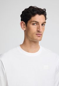 Vit långärmad skjorta i bomull med rund halsringning, med en texturerad lapppatch med orden "POLO CLUB" i ljus färg på bröstet.