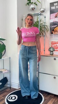 Rožinė trumpa marškinė su "Von Dutch Originals" logotipu, derinamos su šviesiai mėlynomis platėjančiomis džinsomis ir juodais batais ant apvalaus kilimo.