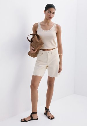 Femme debout en haut beige sans manches et short, tenant un sac marron, portant des sandales noires à lanières et un bracelet en or.