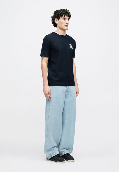 Μπλε βαμβακερό μπλουζάκι με κοντά μανίκια και στρογγυλή λαιμόκοψη, με λογότυπο στο στήθος, συνδυασμένο με ανοιχτό μπλε τζιν παντελόνι φαρδιάς γραμμής.