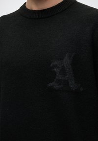 Gros plan d'un pull en maille noir avec une lettre sombre subtilement texturée "A" au niveau de la poitrine.
