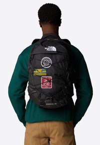 Black backpack with multiple patches, featuring a drawstring design, adjustable straps, and front bungee cord for extra storage.