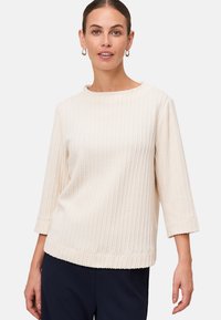 Femme portant un pull en tricot texturé crème avec des manches 3/4 et un pantalon marine, se tenant devant un fond gris clair uni.