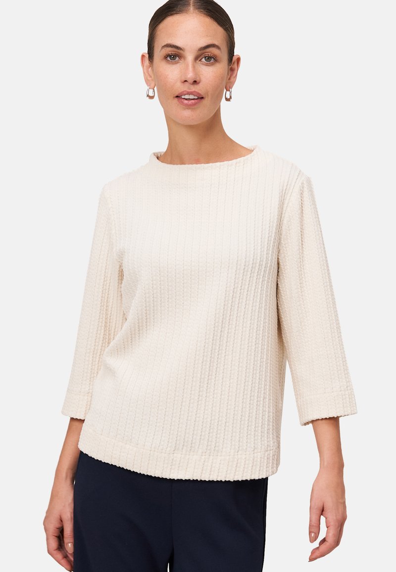 Femme portant un pull en tricot texturé crème avec des manches 3/4 et un pantalon marine, se tenant devant un fond gris clair uni.