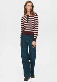 Frau, die einen gestreiften, kastanienbraunen und weißen Pullover mit korallefarbigem Kragen trägt, dazu weite, dunkelblaue Jeans und braune Schuhe, steht vor einem weißen Hintergrund.