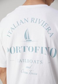 Balta medvilnė marškinėliai su šviesiai mėlynu spaudiniu nugaroje, kuriame yra tekstas "ITALIAN RIVIERA PORTOFINO SAILBOATS and Ocean Tours" ir burlaivio grafika.