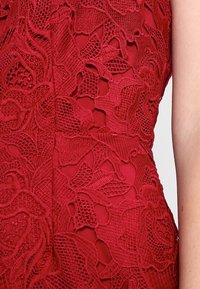 Röd spetsklänning med blommönster, texturerad tyg och figurpassande design. Funktioner inkluderar intrikata detaljer och ett slätt underlager.