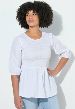 Kvinde med krøllet hår iført en hvid smocket bluse med puffede ærmer og lyseblå jeans, stående med den ene hånd på hoften.