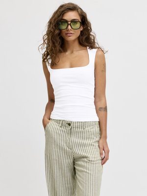 Woman with curly hair wearing green-tinted sunglasses, a white square-neck tank top, and beige striped pants with hands in pockets.