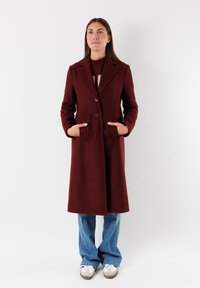 Cappotto di lana bordeaux con rever a soffietto, due tasche frontali, chiusura a doppio bottone e vestibilità sartoriale, abbinato a jeans chiari a zampa d'elefante e sneakers.
