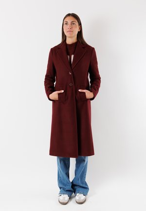 Cappotto di lana bordeaux con rever a soffietto, due tasche frontali, chiusura a doppio bottone e vestibilità sartoriale, abbinato a jeans chiari a zampa d'elefante e sneakers.