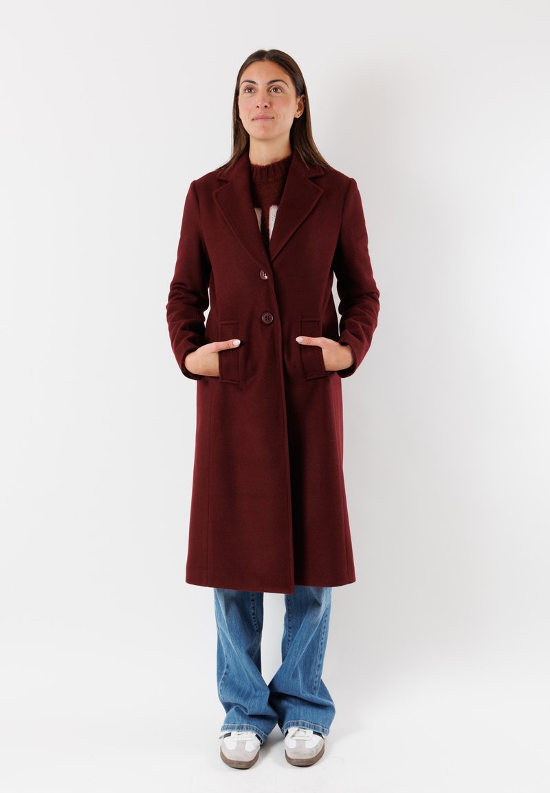 Cappotto di lana bordeaux con rever a soffietto, due tasche frontali, chiusura a doppio bottone e vestibilità sartoriale, abbinato a jeans chiari a zampa d'elefante e sneakers.