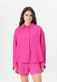 Camicia button-up rosa in tessuto texturizzato con taschino e colletto, abbinata a pantaloni corti rosa all'uncinetto con coulisse.
