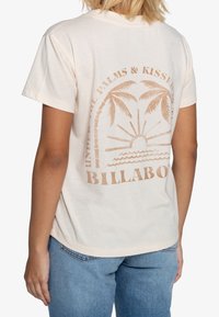 Osoba v světle béžovém tričku s potiskem palem, slunce, vln a textem "Billabong" na zádech, kombinované s modrými džínami.