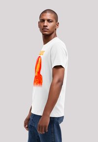 White cotton t-shirt featuring a graphic print with orange and red tones of a person, and bold yellow text above the image. Short sleeves.