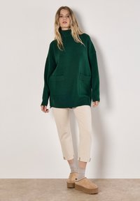 Grüner, übergroßer Rollkragenpullover mit zwei vorderen Taschen, kombiniert mit hellbeigen, knöchellangen Hosen und tanfarbenen Plateauschuhen. Weiche Textur.