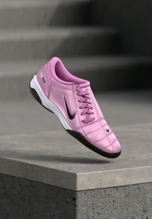 Roze Nike voetbalschoen met zwart swoosh-logo, witte en zwarte zool, tentoongesteld bovenop een betonnen rand met onscherpe trappen op de achtergrond.