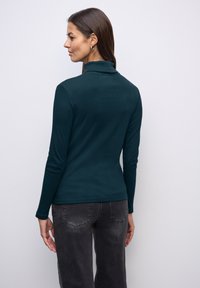 Donkerteal lange-mouwen top met een opstaande kraag, gemaakt van gladde stof. Gecombineerd met grijze jeans, met een slanke pasvorm en eenvoudig ontwerp.