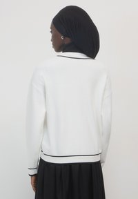 Donna che indossa un maglione bianco con rifiniture nere, una gonna nera e un velo nero, vista di spalle su uno sfondo semplice.