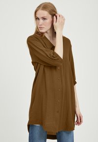 Chemise marron à boutons avec col, manches mi-longues et coupe décontractée. Le tissu semble léger avec une texture douce.