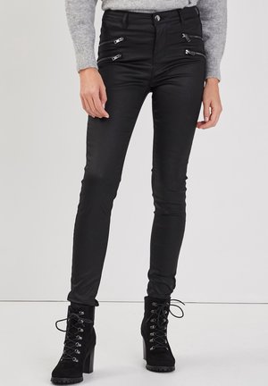 Pantalon noir slim avec quatre fermetures éclair à l'avant, associé à des bottes à talons noires à lacets, porté par une personne portant un pull gris clair.